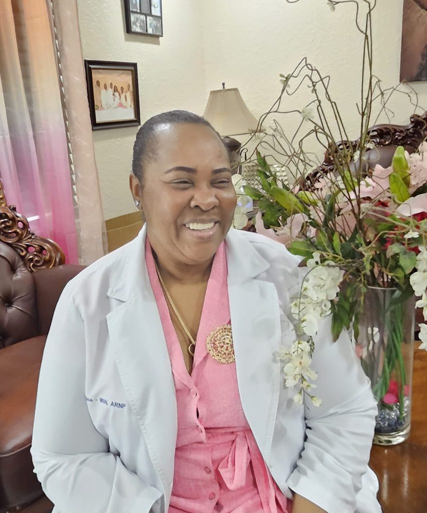 A smiling woman in a white coat sits in a well-decorated room with a floral arrangement.