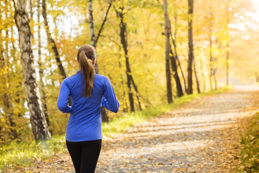 A woman jogs along a trail surrounded by vibrant autumn foliage and trees. The path is covered in fallen leaves.